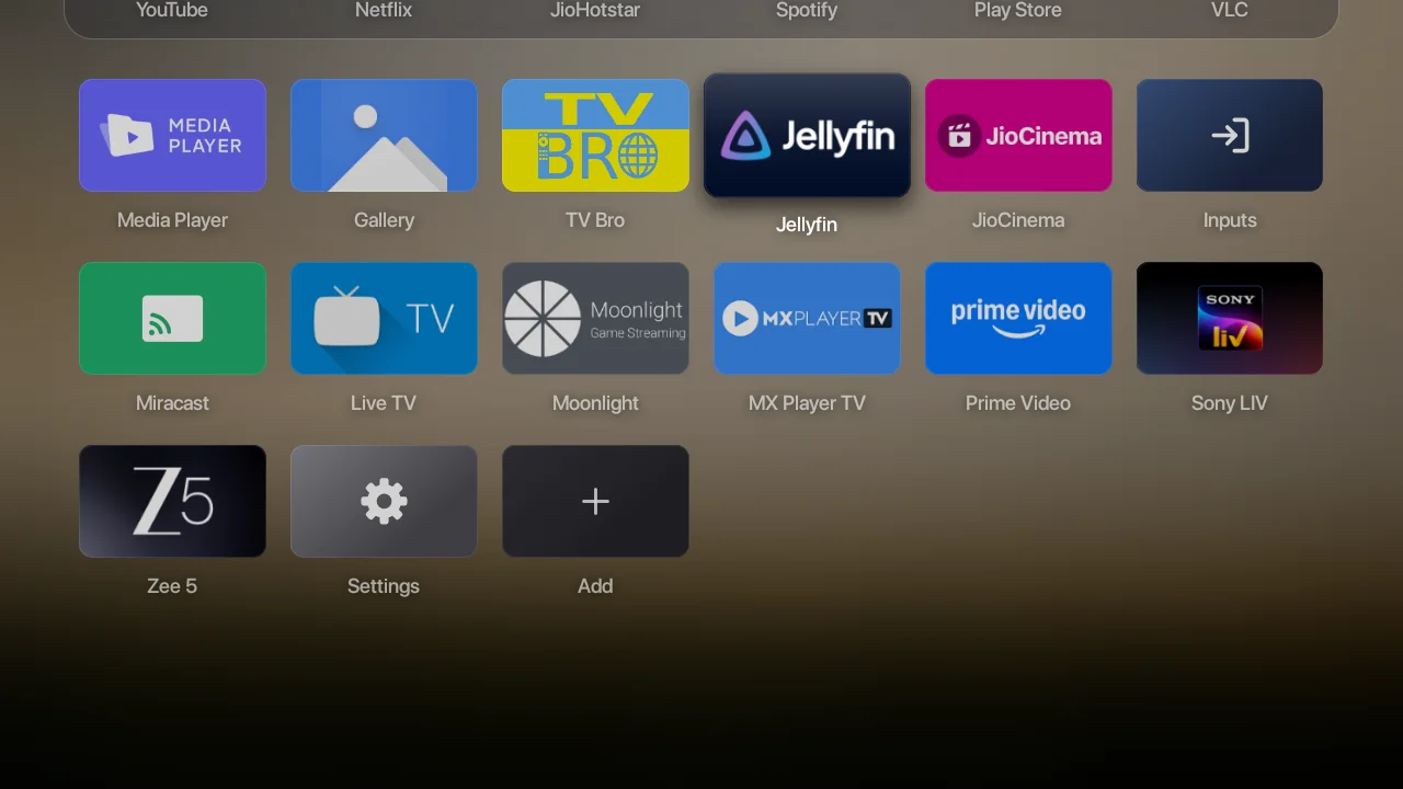 Bare's full app grid on my TV: Media Player, Gallery, TV Bro, Jellyfin, JioCinema, Prime Video, Sony LIV, Zee 5 and more, with names, over a frosted, darkened copy of the photo.