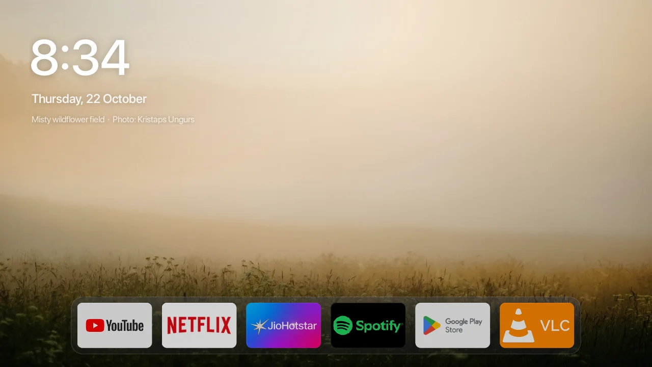 Bare on my TV: the time and date over a misty meadow at sunrise, the photo's caption under the date, and YouTube, Netflix, JioHotstar, Spotify, Play Store and VLC in a small glass dock along the bottom.