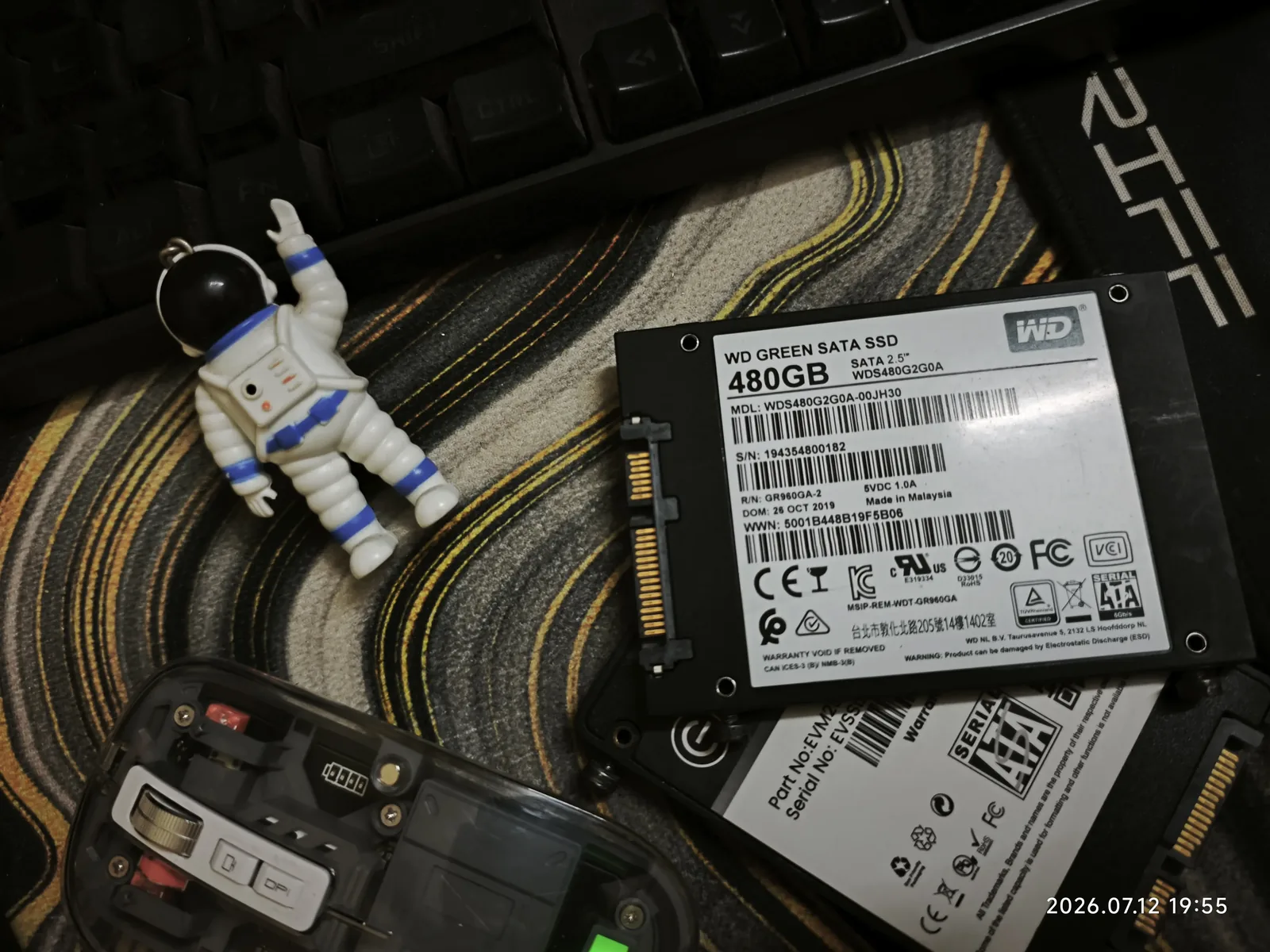 A WD Green 480GB SATA SSD pulled out of the desktop, next to a mechanical keyboard, a small astronaut keychain, and a partially disassembled remote, mid desk teardown.